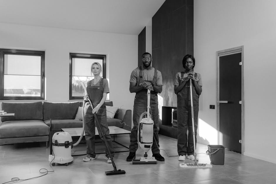 Three professional cleaners from Kennington Carpet Cleaners are depicted performing surface cleaning and deep cleaning in a modern living room. The room features a large, L-shaped fabric sofa marked by a smooth, dust-free surface, positioned against a wall with two large windows allowing natural light to illuminate the space. The floor is covered with polished wooden material, appearing clean and shiny, indicating recent sanitisation. One cleaner, standing at the left, is operating a portable vacuum cleaner with a long hose, ensuring thorough removal of dust from the laminate flooring near a glass coffee table. In the center, another cleaner uses a handheld spray bottle and cloth, likely for surface disinfection or spot cleaning. The third cleaner on the right is mopping the floor with a long mop, with a bucket placed nearby, suggesting ongoing floor maintenance. All three individuals are wearing work uniforms and appear focused on their tasks, contributing to a hygienic, well-maintained environment achieved by professional domestic cleaning services. The scene emphasizes surface cleaning and sanitisation processes in a bright, tidy residential setting, indicative of the quality service provided by Kennington Carpet Cleaners, aimed at maintaining cleanliness and hygiene in homes.