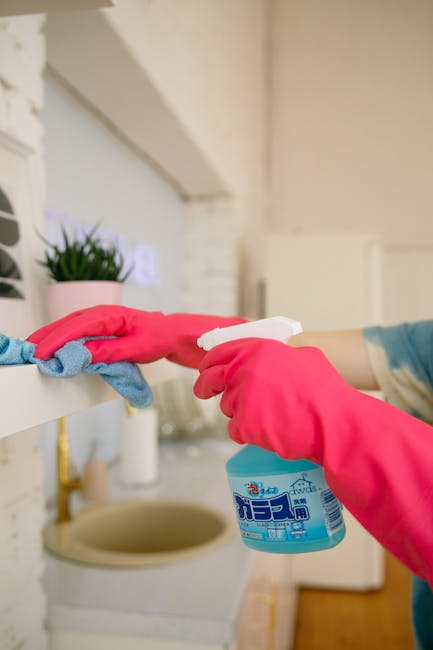 A female cleaning professional with light skin and shoulder-length brown hair, wearing a yellow uniform and a badge, stands in a well-lit living room holding a spray bottle in her right hand and a blue microfiber cloth in her left. The room features a beige sofa with cushions, a wooden side table with decorative items, and a large window allowing natural light to illuminate the space. The surfaces visible include a glass display cabinet, a wooden table, and a tiled fireplace mantel, all appearing clean and well-maintained. The setting highlights a tidy, hygienic environment suitable for residential deep cleaning services, and the professional from Kennington Carpet Cleaners is prepared for surface cleaning or sanitisation tasks as part of their domestic cleaning offerings, aligned with the topic of accurate cleaning quotes and instant estimates in Kennington SE11.