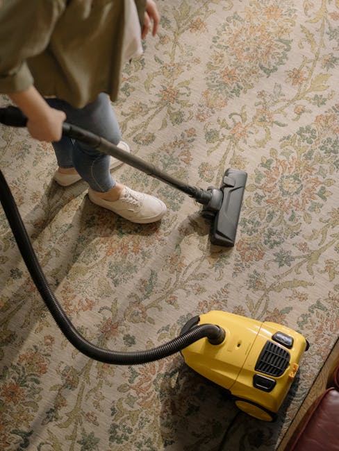 A person wearing beige shoes and blue jeans is operating a yellow vacuum cleaner with a black hose, cleaning an ornate, patterned area rug made of floral motifs in muted colors. The room's lighting highlights the clean, shiny surface of the rug, which appears freshly vacuumed and well-maintained. The vacuum cleaner's controls and hose are clearly visible, situated on a wooden floor adjacent to the rug. This scene exemplifies professional carpet cleaning services provided by Kennington Carpet Cleaners as part of their deep cleaning and surface sanitisation offerings near Kennington Park.