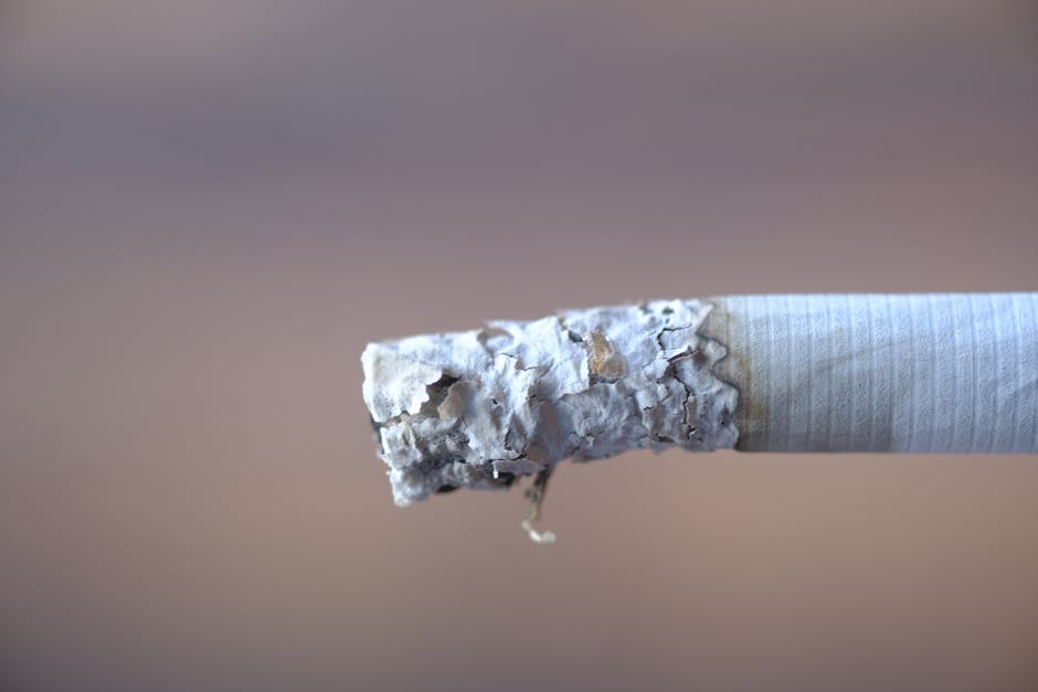 Close-up of a burnt-out cigarette stub with a crumpled, white ash tip and a partially flattened filter, resting horizontally on a neutral, blurred background. The cigarette's paper wrapper appears sli