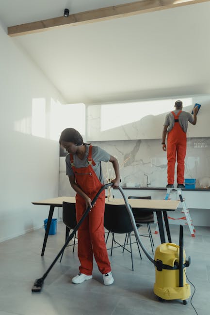 Two professional cleaners from Kennington Carpet Cleaners are performing surface cleaning and deep cleaning activities inside a modern residential kitchen. One cleaner, positioned in the foreground, is using a vacuum cleaner on the light gray wooden or laminate floor, creating a dust-free and hygienic environment. The other cleaner, standing on a small step ladder, is wiping or polishing the white marble backsplash with a cloth. The kitchen features a clean, minimalistic design with a white ceiling, natural light flooding through large windows, and a central table with black chairs, indicating a bright and well-maintained space. Visible cleaning tools and a yellow pressure washer placed nearby highlight the emphasis on thorough sanitation, aligning with professional domestic cleaning standards promoted by Kennington Carpet Cleaners, who emphasize transparent pricing without hidden charges when booking cleaning services.