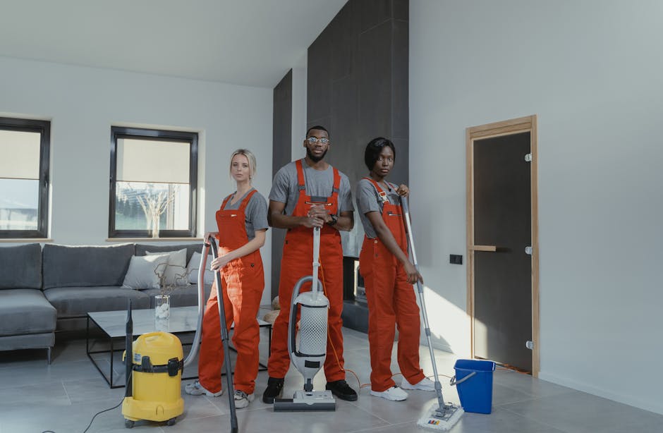 Two professional cleaners from Kennington Carpet Cleaners are performing surface cleaning and deep cleaning activities inside a modern residential kitchen. One cleaner, positioned in the foreground, is using a vacuum cleaner on the light gray wooden or laminate floor, creating a dust-free and hygienic environment. The other cleaner, standing on a small step ladder, is wiping or polishing the white marble backsplash with a cloth. The kitchen features a clean, minimalistic design with a white ceiling, natural light flooding through large windows, and a central table with black chairs, indicating a bright and well-maintained space. Visible cleaning tools and a yellow pressure washer placed nearby highlight the emphasis on thorough sanitation, aligning with professional domestic cleaning standards promoted by Kennington Carpet Cleaners, who emphasize transparent pricing without hidden charges when booking cleaning services.