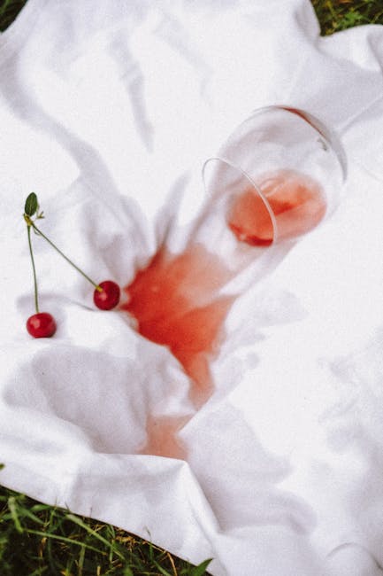 A tall, clear glass tipped over on a white fabric surface, with a small amount of pinkish-red liquid spilled around it, suggesting a red wine stain. Two cherries with their stems attached are also resting on the fabric, near the glass. The scene appears to be outdoors, with natural sunlight illuminating the fabric and objects, highlighting the clean and fresh appearance. This image relates to surface cleaning and stain removal, aligning with the topic of residential and commercial cleaning as discussed by Kennington Carpet Cleaners in their page on red wine stain and carpet cleaning in Kennington.