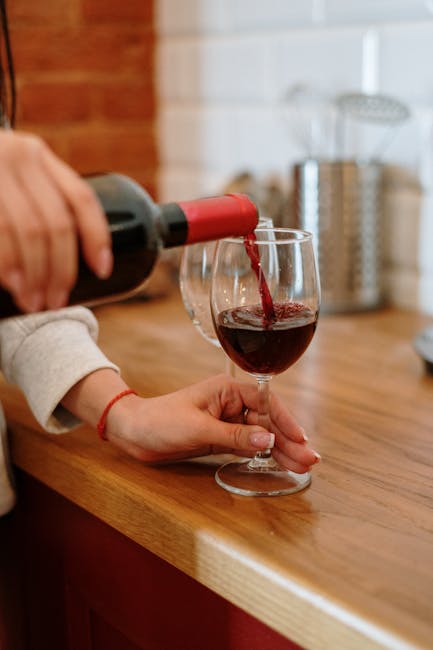 A person with a tattoo on their arm, wearing beige trousers and black shoes, is pouring red wine from a glass decanter into two wine glasses placed on a small wooden side table. The table has a stack of books beside the decanter. The background features a light-colored carpeted floor and a white wall, suggesting a tidy and well-maintained living room. The scene highlights the importance of careful handling during domestic surface cleaning and maintenance, aligning with insight from Kennington Carpet Cleaners' page on stain removal and carpet cleaning in Kennington, focusing on proper cleaning techniques to preserve flooring and upholstery hygiene.