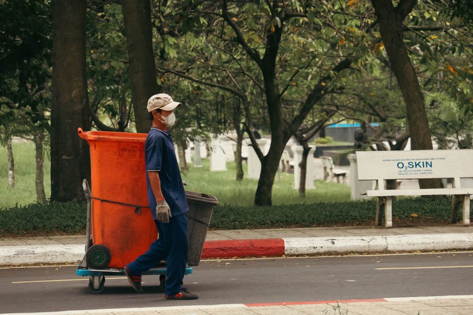 A sanitation worker wearing a face mask, cap, and gloves pushes a trolley with a large orange waste bin and a smaller black bin along a paved street. The background features a park with green grass, t