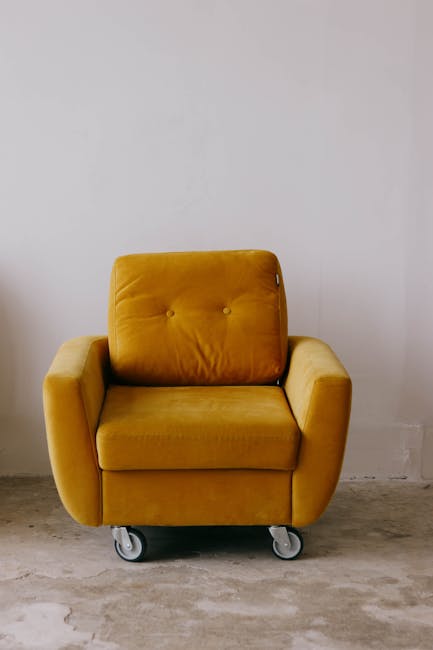A mustard yellow upholstered armchair with a modern design, featuring a cushioned backrest with two buttons, wide armrests, and a thick seat cushion, situated on a concrete floor against a plain white wall. The chair is mounted on small caster wheels, indicating ease of movement. The room appears well-lit, and the overall setting reflects a clean, minimalist interior. This image highlights the furniture piece that could benefit from professional cleaning and sanitisation services offered by Kennington Carpet Cleaners as part of surface cleaning and deep cleaning for residential or commercial spaces, in accordance with Lambeth Council rules for upholstery cleaning in Kennington.