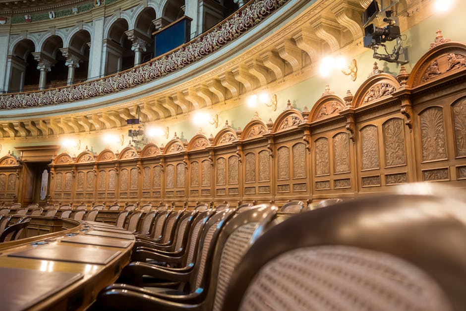 Interior view of a historic legislative chamber featuring polished wooden surfaces, ornate wood paneling with decorative carvings, and matching wooden chairs with woven cane seats arranged around a long, curved wooden table. The room is illuminated by warm wall-mounted sconces, highlighting the craftsmanship of the woodwork on the walls and ceiling. The space appears exceptionally clean and well-maintained, emphasizing surface hygiene and deep cleaning. Visible tools or cleaning products are not present, but the pristine condition suggests recent professional sanitisation and maintenance by Kennington Carpet Cleaners to comply with Lambeth Council rules for upholstery cleaning in Kennington. The lighting creates a welcoming atmosphere suitable for formal proceedings, with attention to surface cleanliness and overall hygiene.