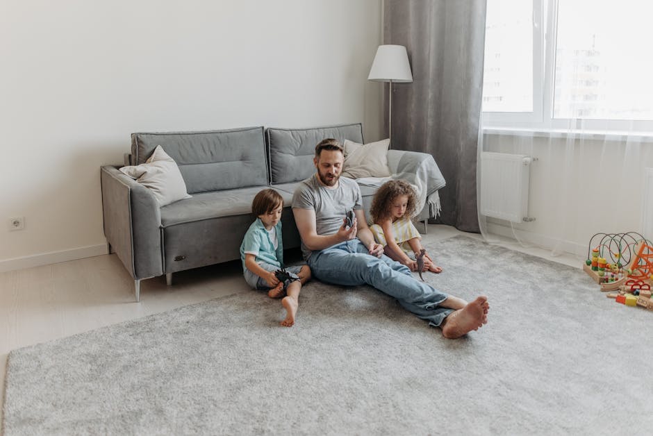A man sitting on a light-colored, plush carpet in a bright living room, surrounded by two young children. The man and children are barefoot, with the man holding a remote control and the children playing with toys. The room features a modern, light gray sofa with white cushions, standing against a plain white wall. A tall white floor lamp is positioned beside the window, which allows natural light to fill the space. The window has sheer white curtains, and a radiator is installed beneath it. To the right, a small wooden toy bead maze typical of children's play areas is visible on the floor. The carpet and the overall room appear clean and well-maintained, consistent with professional deep cleaning or sanitation standards related to surface cleaning and domestic cleaning, as promoted by Kennington Carpet Cleaners.
