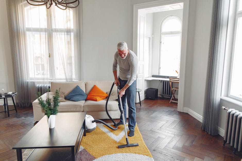 A man with gray hair and a gray sweater vacuuming a living room with a black vacuum cleaner, which is placed on a multicolored area rug with yellow, pink, and white patterns. The room features a white sofa with colorful pillows, including orange and blue, positioned against large windows with sheer curtains. The hardwood floor is visible around the rug, and a small black side table with a water bottle is situated next to the sofa. In the background, there is a white radiator beneath one window, a small desk with a chair in the corner, and a window seat with its window letting in natural light. The room appears well-maintained, clean, and bright, emphasizing professional surface cleaning and deep cleaning activities supported by Kennington Carpet Cleaners.