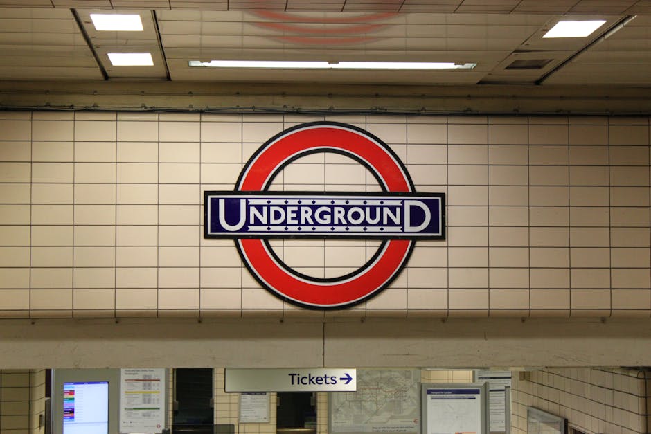Close-up of a London Underground station sign on a tiled wall, displaying the iconic red circle and blue bar with the word 'UNDERGROUND' printed in white letters. Below the sign, there is a white directional sign pointing towards ticketing areas and a partial view of ticket machines and station information boards, all situated in a clean, well-lit underground corridor. The surface appears hygienic and well-maintained, reflecting the thorough surface cleaning and sanitisation services provided by Kennington Carpet Cleaners, as referenced on the webpage titled 'Urgent same day cleaning for Kennington tube and Oval area' on their website.