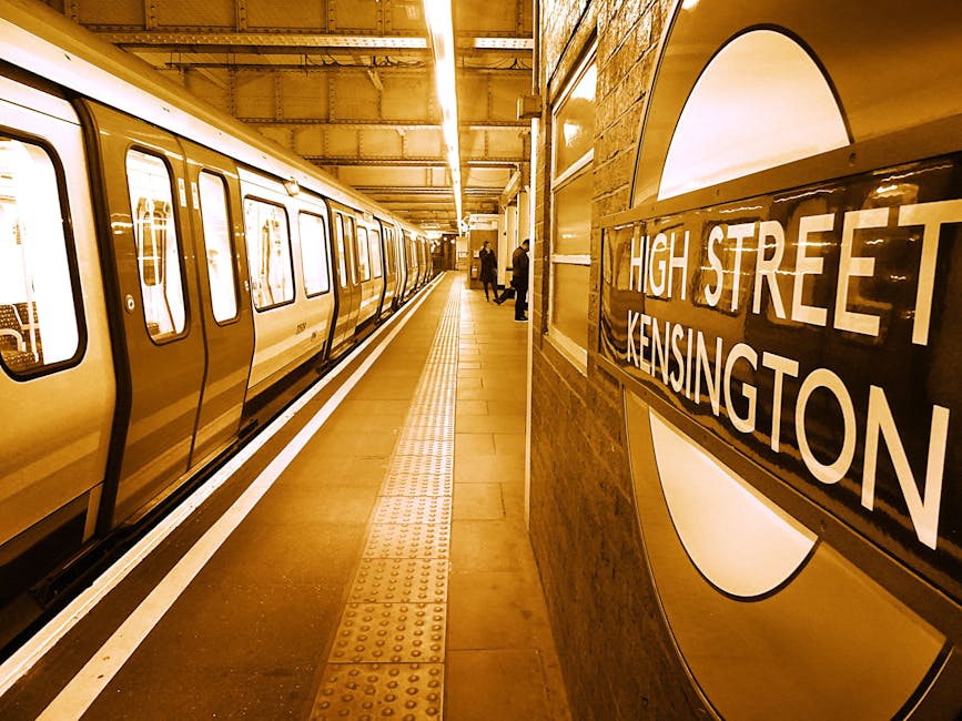 Close-up of a London Underground station sign on a tiled wall, displaying the iconic red circle and blue bar with the word 'UNDERGROUND' printed in white letters. Below the sign, there is a white directional sign pointing towards ticketing areas and a partial view of ticket machines and station information boards, all situated in a clean, well-lit underground corridor. The surface appears hygienic and well-maintained, reflecting the thorough surface cleaning and sanitisation services provided by Kennington Carpet Cleaners, as referenced on the webpage titled 'Urgent same day cleaning for Kennington tube and Oval area' on their website.